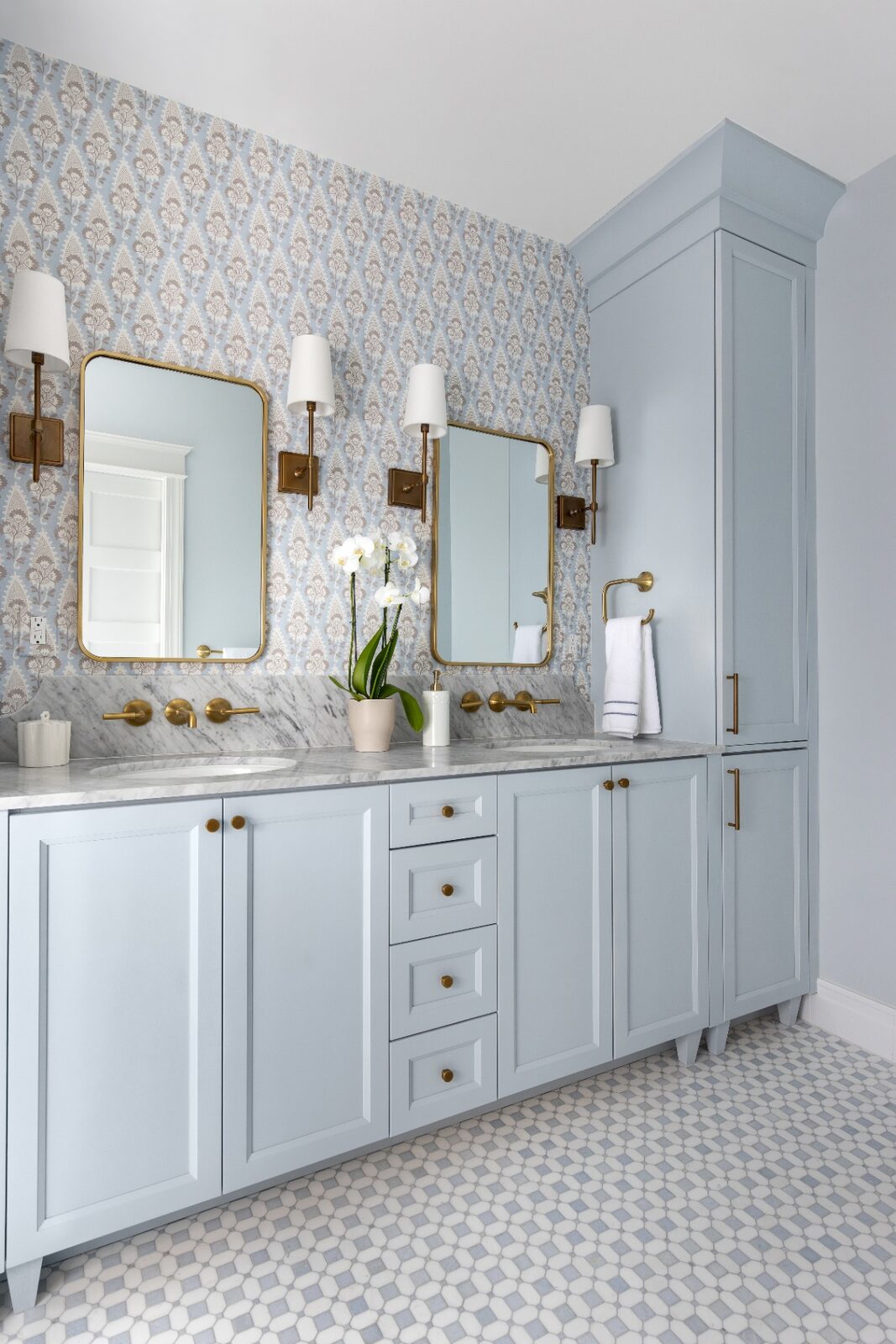 Soft blue double-vanity primary bath with marble counters, brass sconces and patterned wallpaper