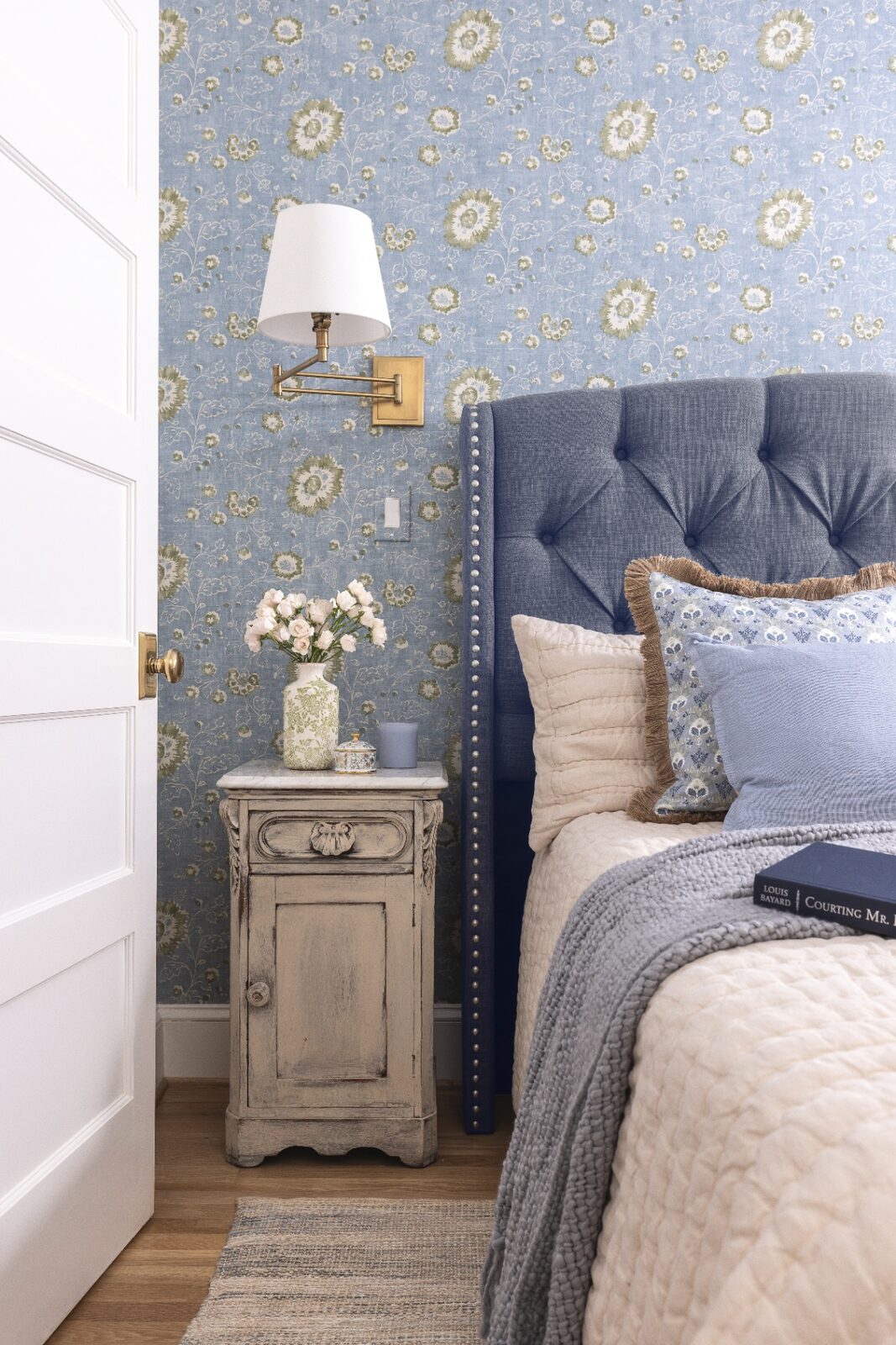 Guest bedroom with chambray-blue wallpaper, tufted headboard and antique nightstand